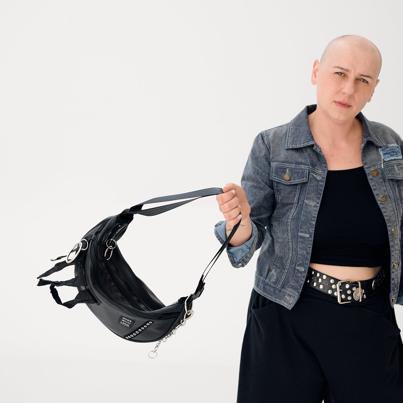 Eine Frau in Jeansjacke hält eine schwarze Moonwalker-Tasche aus Leder am Riemen, mit Ringen und Kette an der Naht